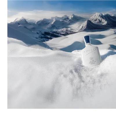 A white perfume bottle with a dark blue cap is nestled in glistening white snow, with blurry snow-covered mountains in the background, and the bottle displays text including CREED, SILVER MOUNTAIN WATER, DE PERE EN FILS DEPUIS 1760, FROM FATHER TO SON SINCE 1760, 100 ML, 3.33 FL. OZ.