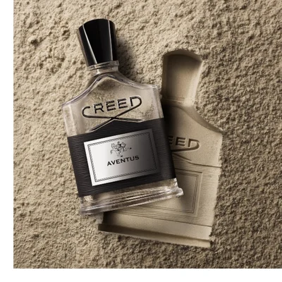 A Creed Aventus perfume bottle is shown in front of a pineapple with the text CREED AVENTUS visible on the bottle.