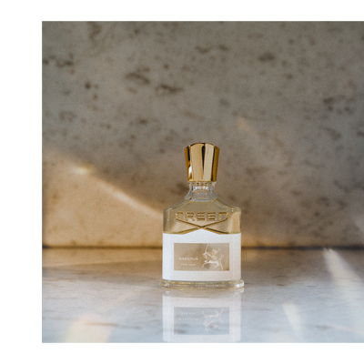 A clear glass bottle of Creed Aventus Cologne perfume with a silver cap rests on a marble surface, featuring the logo CREED and label text AVENTUS COLOGNE