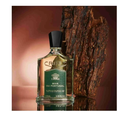 A bottle of Creed perfume is shown next to a piece of bark, with the label reading CREED BOIS DU PORTUGAL DE PERE EN FILS DEPUIS 1760 FROM FATHER TO SON SINCE 1760 100ML 3.33 FL. OZ