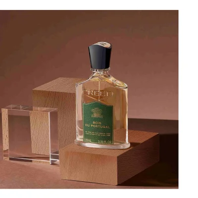 A Creed perfume bottle with a green label sits on a wooden block displaying the text CREED BOIS DU PORTUGAL DE PERE EN FILS DEPUIS 1760 FROM FATHER TO SON SINCE 1760 100ML 3.33 FL. OZ.