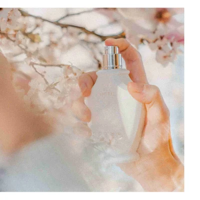 A hand holds a white perfume bottle with the text LOVE IN WHITE in front of blurred pink and white flowers.