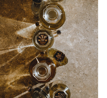Overhead view of several perfume bottles on a stone surface with diagonal light patterns, one bottle shows the text CREED 1760.