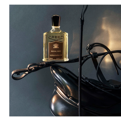 A Creed Royal Oud perfume bottle is displayed on black leather equestrian gear including a saddle and bridle, with visible text CREED ROYAL OUD DE PERE EN FILS DEPUIS 1760 FROM FATHER TO SON SINCE 1760 100ML 3.33 FL. OZ.