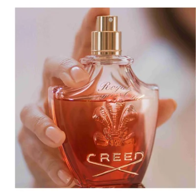 Hand holding a glass perfume bottle with amber liquid and a gold cap featuring the text Royal Princess Oud and CREED.