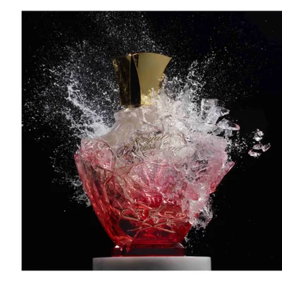 A red and clear perfume bottle with a gold cap is surrounded by a splash of water and ice fragments against a black background and shows the text Royal Princess Oud and CREED.