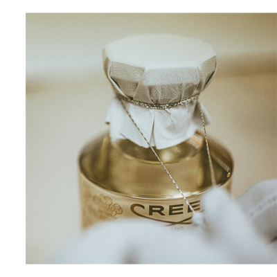 Close-up of a golden bottle, likely perfume, with a cloth-covered cap tied with a string, showing part of the bottles label with text CREED 1760.