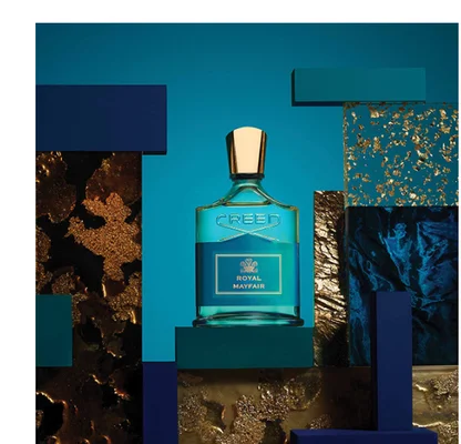 A Creed Royal Mayfair perfume bottle is displayed on a teal block surrounded by abstract blocks in blue, teal, gold texture, and dark blue texture against a blue background, with text reading CREED ROYAL MAYFAIR.