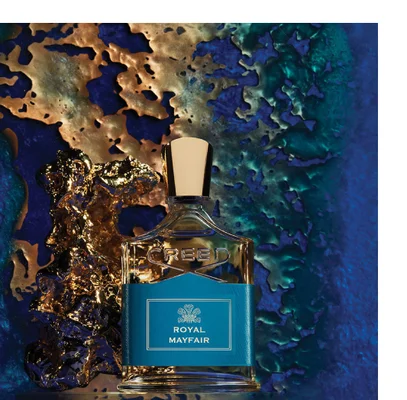 A perfume bottle with a blue label stands next to a gold object against a textured blue and gold background with text reading CREED ROYAL MAYFAIR.