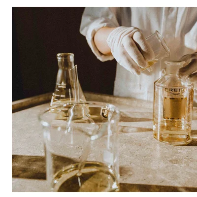 Person wearing gloves pours liquid from a beaker into a glass bottle with the label CREED Millesime Imperial 1760.