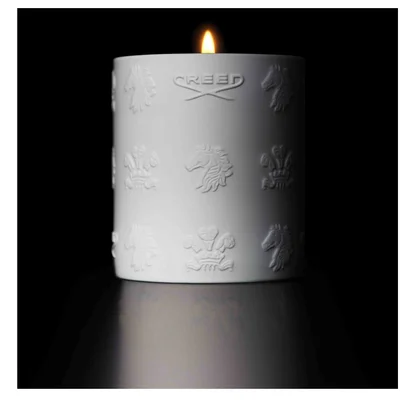 A lit white candle with embossed designs and the text CREED sits on a reflective surface in a dark setting.