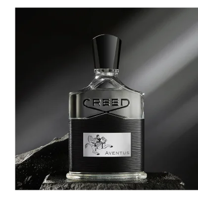 A bottle of Creed Aventus perfume with a black ribbed base, silver band, and a label showing a rider on a horse, sits on a dark surface displaying the text CREED and AVENTUS.