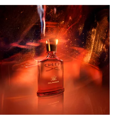 A red perfume bottle with a gold cap is shown against a fiery orange background with gold sparkles, with the text CREED DELPHINUS visible on the bottle.