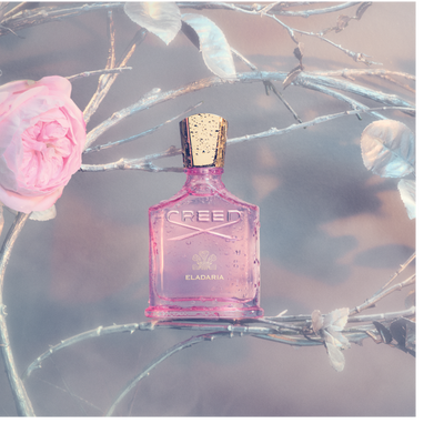 A pink Creed Eladaria perfume bottle covered in water droplets sits among silver branches and a pink rose.