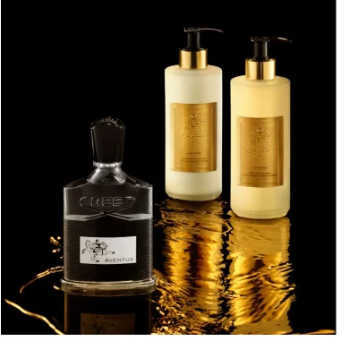 Three luxury fragrance and body products from Creed Aventus, a dark perfume bottle and two pump bottles (one cream, one light yellow), are arranged on a reflective black surface with swirling golden liquid; the dark bottle displays CREED and AVENTUS, the cream pump bottles label reads CREED, 1760, AVENTUS, HAND & BODY LOTION, LOTION MAINS ET CORPS, and the light yellow pump bottles label reads CREED, 1760, AVENTUS, HAND & BODY WASH, SAVON LIQUIDE MAINS ET CORPS.