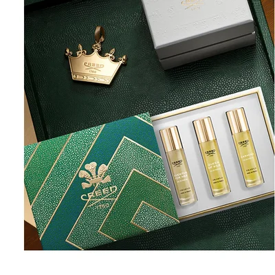 An open green and gold geometric patterned gift box contains three small perfume rollerballs labeled Aventus For Her, Queen of Silk, and Carmina, displayed on a dark green textured surface alongside a gold crown charm and a white box; visible text includes Creed 1760 on the gift box, crown, and each perfume bottle, with each bottles full label reading Creed 1760 Aventus For Her Eau de Parfum 10 ML 0.33 FL.OZ. MADE IN FRANCE, Creed 1760 Queen of Silk Eau de Parfum 10 ML 0.33 FL.OZ. MADE IN FRANCE, and Creed 1760 Carmina Eau de Parfum 10 ML 0.33 FL.OZ. MADE IN FRANCE.