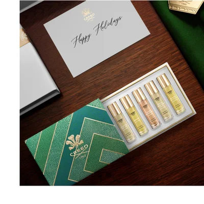 An open green and gold geometric patterned gift box contains three small perfume rollerballs labeled Aventus For Her, Queen of Silk, and Carmina, displayed on a dark green textured surface alongside a gold crown charm and a white box; visible text includes Creed 1760 on the gift box, crown, and each perfume bottle, with each bottles full label reading Creed 1760 Aventus For Her Eau de Parfum 10 ML 0.33 FL.OZ. MADE IN FRANCE, Creed 1760 Queen of Silk Eau de Parfum 10 ML 0.33 FL.OZ. MADE IN FRANCE, and Creed 1760 Carmina Eau de Parfum 10 ML 0.33 FL.OZ. MADE IN FRANCE.