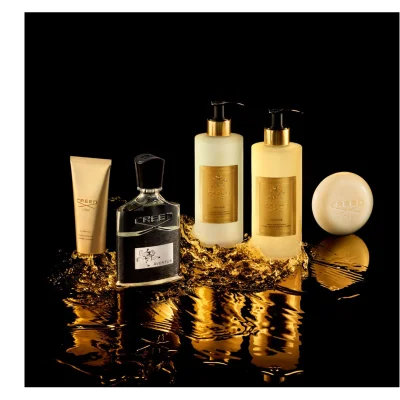 Five Creed Aventus products, including a gold hand cream tube, a black perfume bottle, two pump bottles for hand and body lotion and wash, and a round soap bar, are displayed amidst golden splashing liquid on a dark surface, featuring text Creed 1760, Aventus, Hand Cream, Crème Mains, Hand & Body Lotion, Lotion Mains & Corps, Hand & Body Wash, and Lavant Liquide Mains & Corps.