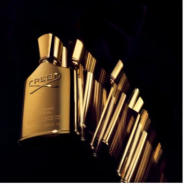 A shiny gold Creed Millésime Impérial perfume bottle is angled against a dark background with a trailing light effect, displaying the text CREED, MILLÉSIME IMPERIAL, DE PÈRE EN FILS DEPUIS 1760, FROM FATHER TO SON SINCE 1760, 100 ML, and 3.33 FL. OZ.