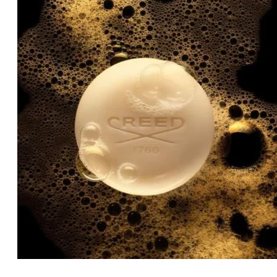 A round, light-colored bar of soap rests in golden-brown foam with many bubbles on a dark background, featuring the embossed text CREED 1760.