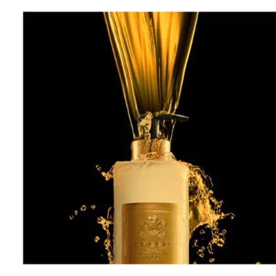 A light-colored bottle with a gold label has golden liquid pouring into it from above, creating splashes, with the text CREED 1760 visible on the label.