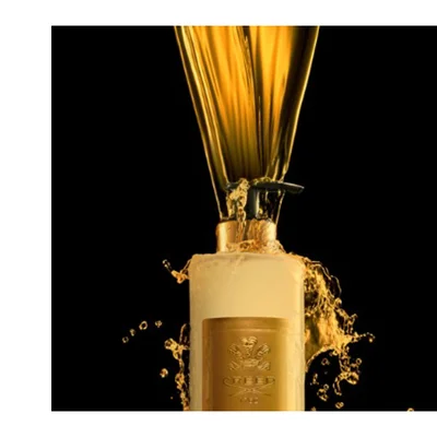 A light-colored Creed bottle with a gold label has golden liquid pouring into its dispenser pump, creating splashes, with the visible text Creed 1760.