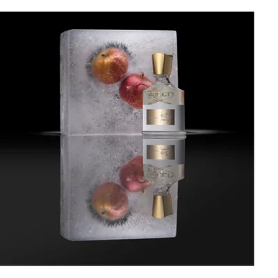 A clear glass perfume bottle with a gold cap and visible text Creed Aventus stands on a reflective dark surface beside a block of ice containing three red and yellow apples.