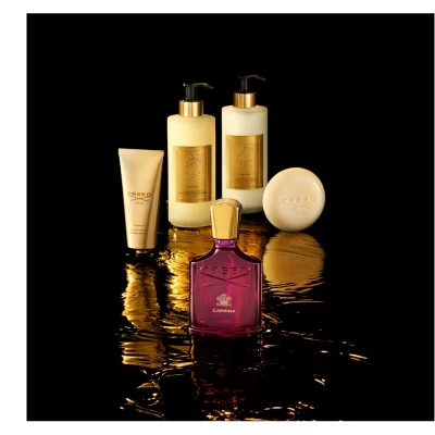 A collection of five luxurious Creed beauty products including a plum-colored Carmina perfume bottle, a cream hand cream tube, two cream pump-top bottles for hand and body wash and lotion, and a cream oval bar of soap, are artfully arranged on a dark, reflective surface with shimmering gold light, featuring the text CREED 1760 CARMINA HAND CREAM CRÈME MAINS HAND & BODY WASH SAVON LIQUIDE MAINS & CORPS HAND & BODY LOTION LAIT MAINS & CORPS.