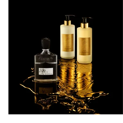 Creed Aventus bottle with matching lotion and wash, reflected in gold liquid on a black background.